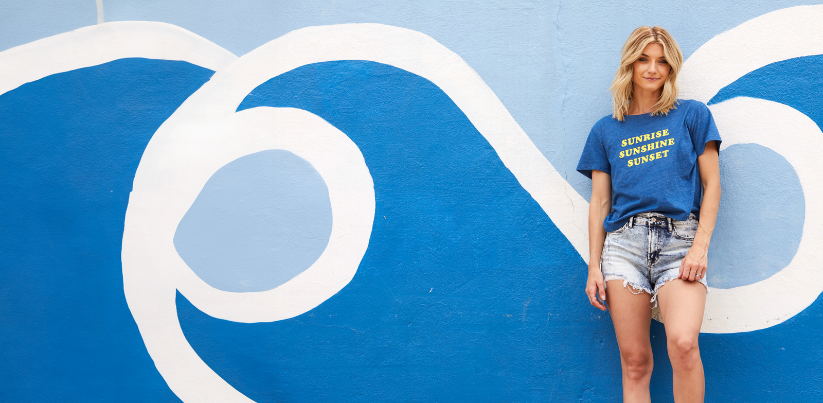 Banner of a blonde woman leaning against a blue wall with white waves painted on it.