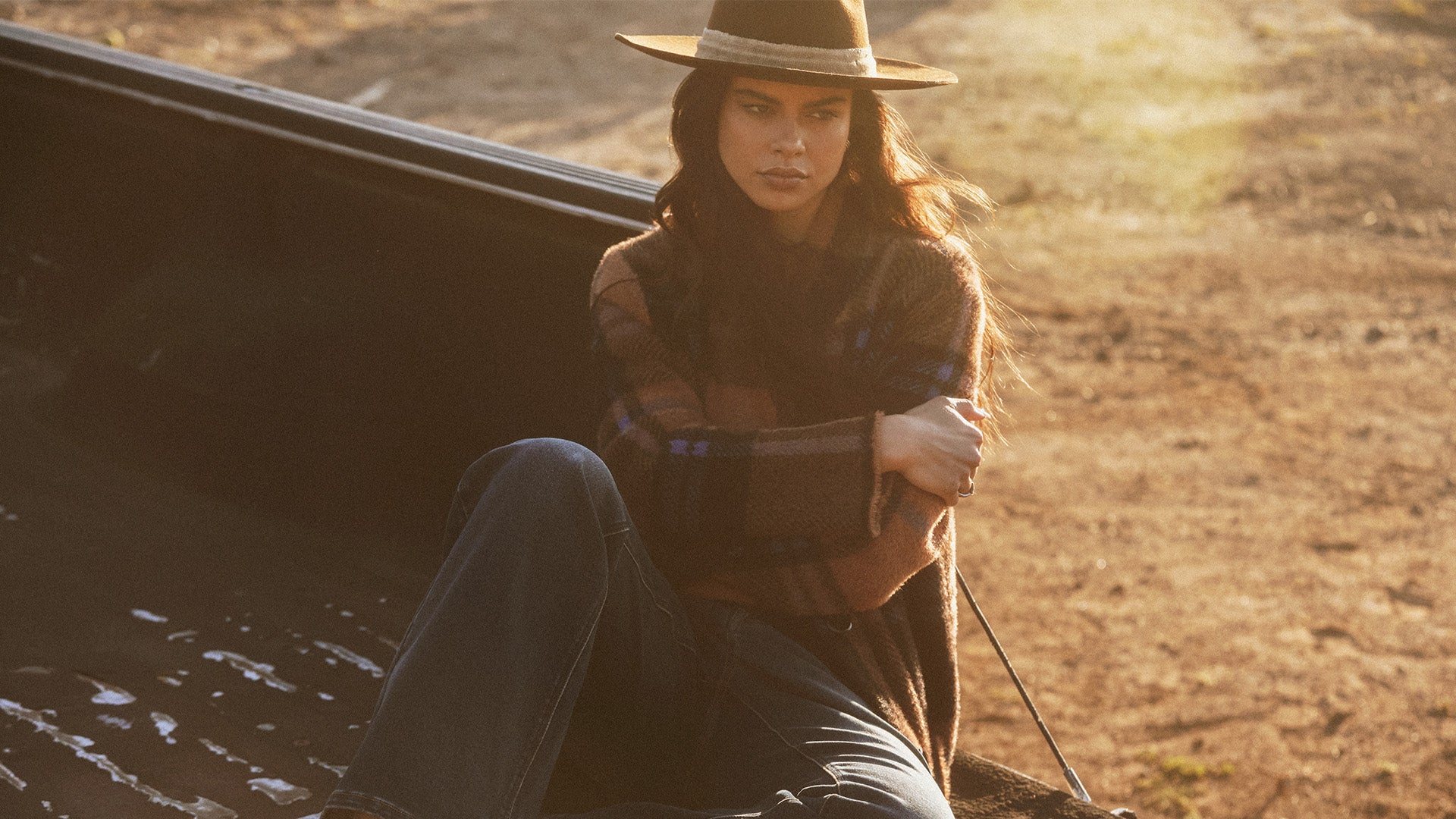 Woman sitting in a truck bed wearing a hat and plaid shirt with a sunset in the background